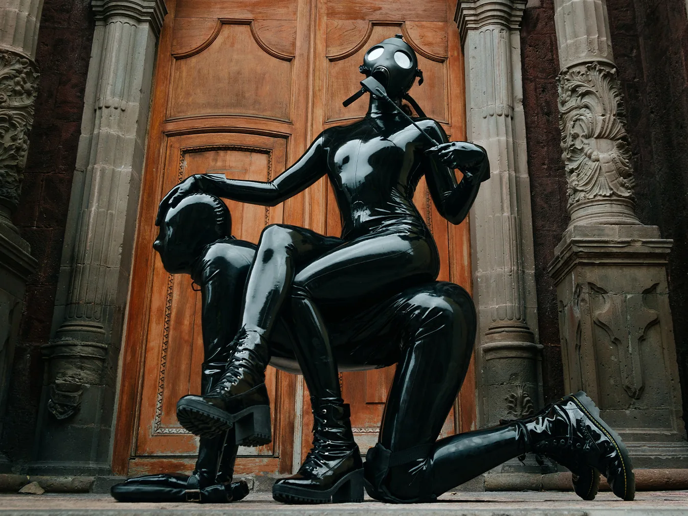 Zwei Personen komplett in schwarzem Latex mit Gasmaske vor einer historischen Holztür