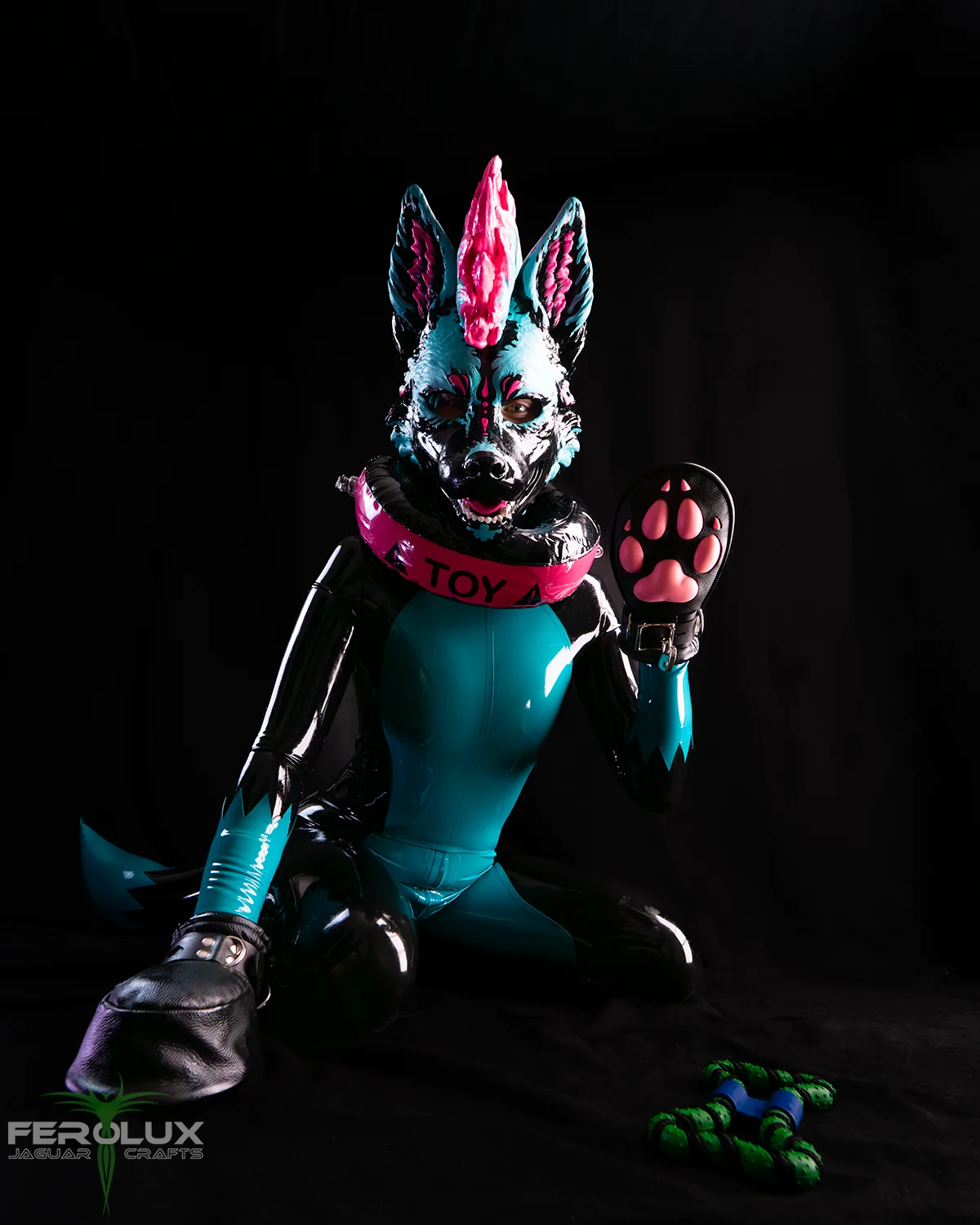 Latex-Pup im türkis-schwarzen Suit zeigt eine rosa Pfote, mit Toy-Halsband