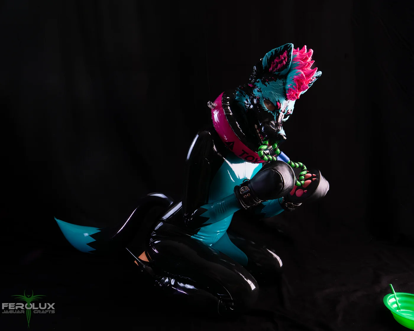 Latex-Pup kniet seitlich und spielt mit den Pfoten, daneben ein Napf